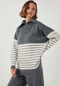 Pulover gri din tricot cu guler cu fermoar, prezentând dungi orizontale crem și gri închis pe mâneci și partea inferioară, cu o croială lejeră.