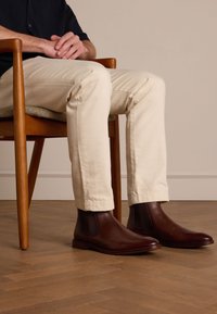Des bottes Chelsea en cuir marron avec des panneaux élastiques sur les côtés et un bout rond, associées à un pantalon beige clair, posées sur un sol en bois.