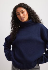 Jūrų mėlynos spalvos golfas, megztas tekstūros, nukritusiais petimi, raukšlėtais rankogaliais ir apatine dalimi, laisvo kirpimo, dėvimas ant pilkų kelnių.