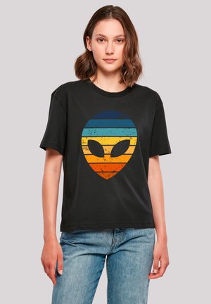 Young woman wearing a black t-shirt with a retro striped alien head design in blue, yellow, and orange, paired with blue jeans.