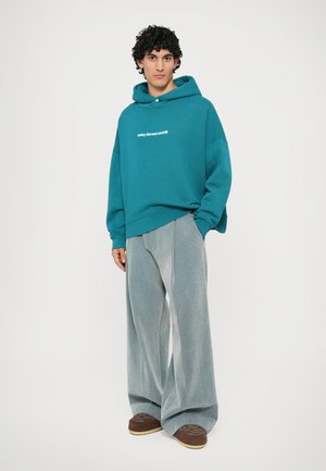 Sweat à capuche surdimensionné teal avec texte blanc, associé à un pantalon large gris clair et des chaussures marron avec une surface texturée.