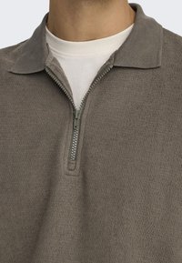Grijze zip-up trui van een gestructureerde stof, met een staande kraag en een zichtbare rits. Gelaagd over een wit T-shirt.