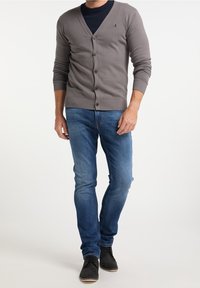 Cardigan grigio con scollo a V, chiusura a bottoni e polsini a coste. Abbinato a jeans blu e scarpe nere. Il tessuto appare morbido e aderente.