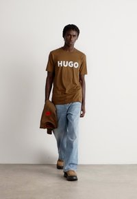 Rudas medvilnės marškinėliai su balta "HUGO" logotipu, derinami su šviesiai mėlynomis džinsinėmis kelnėmis ir rudais švarkais, uždėtais ant rankos. Juodi masyvūs bateliai.