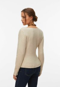 Femme aux cheveux bruns coiffés en chignon, portant un pull beige à manches longues et un jean bleu foncé, posant légèrement de profil sur un fond blanc.