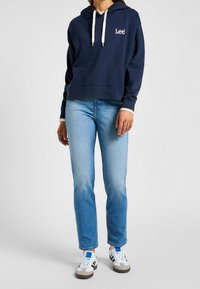 Marinblå huvtröja med vita dragsko och broderad logotyp, kombinerad med ljusblå raka jeans och vita sneakers med detaljer.