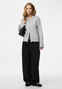 Maglione grigio con cerniera e collo a camicia, abbinato a pantaloni neri larghi e scarpe nere. La modella tiene una borsa nera. Tessuto liscio.