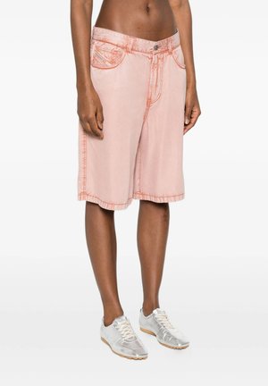 Persona che indossa pantaloncini rosa pallido fino al ginocchio con cuciture arancioni visibili e sneaker argento con lacci, in piedi su sfondo bianco.