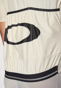 Maillot de cyclisme à manches courtes en crème avec un grand logo noir. Dispose d'une taille élastique avec des rayures horizontales noires et grises contrastées.