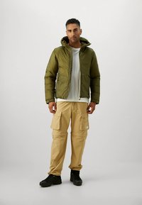 Olivgrön dunjacka med huva, dragkedja framtill och två fickor. Beige cargobyxor med flera fickor. Svarta skor. Enkelt bakgrund.