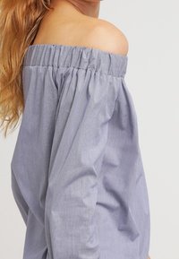 Světle modrá blůza s odkrytými rameny, se staženým výstřihem, dlouhými nažehlenými rukávy a jemnou texturovanou látkou. Minimalistický design a uvolněný střih.