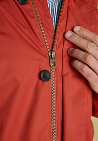 Raudona užtrauktuku užsegama striukė su auksiniu metaliniu užtrauktuku, juodais mygtukais ir šviesia tekstūra. Matoma ranka, esanti netoli užtrauktuko.