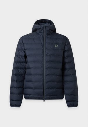 Jeune homme portant un bonnet noir, une doudoune bleu marine, une chemise à carreaux verte, un pantalon noir avec des bandes blanches sur les côtés et des chaussures noires, debout devant un fond uni.