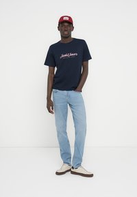 Μπλε ναυτικό βαμβακερό μπλουζάκι με τύπωμα λογότυπου, ανοιχτό μπλε τζιν παντελόνι, μπεζ αθλητικά παπούτσια και μπορντό καπέλο που φοριέται ελαφρώς γέρικο.