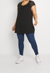 Top negro sin mangas con mangas de encaje, con un pliegue en el frente; combinado con jeans ajustados azules y zapatillas altas blancas.