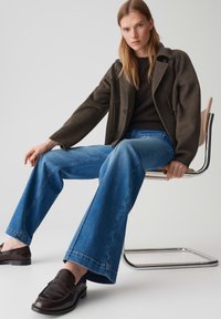 Braune Wollmischungjacke, dunkler Pullover, hellblaue Schlagjeans und schwarze Slipper. Sitzt auf einem Stuhl mit einem Chromrahmen, minimaler Hintergrund.