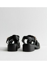 Sandali con tacco nero e design a fascia larga, caratterizzati da ritagli circolari e un accento in oro sulla cinghia alla caviglia. Tacco piatto.