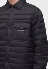 Veste matelassée noire avec boutons-pression et deux poches poitrine, avec le logo blanc "Armani Exchange Milano/New York" sur la poche gauche.