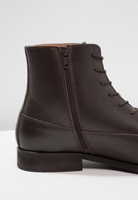 Tmavě hnědá kožená kotníková bota s hladkým povrchem, se zipem na boku a kontrastními tkaničkami. Nízký podpatek pro pohodlí.
