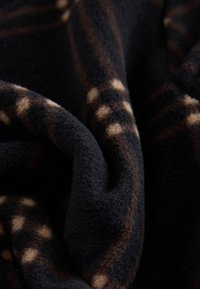 Fleece-tæppe i sort med brunt ternmønster og cremefarvede prikker, med en blød, struktureret overflade og krøllede kanter.