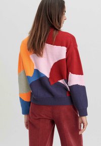 Pull en tricot coloré avec un design en patchwork rouge, orange, rose, bleu et bleu marine. Tissu texturé avec des poignets et un ourlet côtelés.