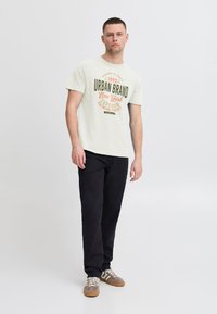 Svetlo zelena bombažna majica s pogumnimi napisi "URBAN BRAND" v oranžni in rjavi barvi, v kombinaciji z črnimi hlačami in rjavimi čevlji z belimi detajli.