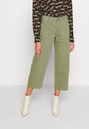 Personne portant un pantalon olive large et court, un haut noir à motif animal beige, et des bottines blanches, debout devant un fond uni.