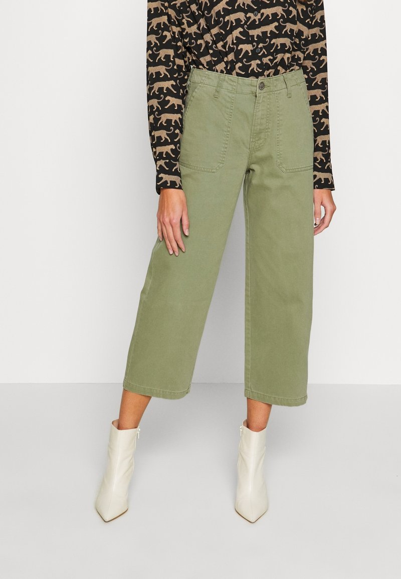 Personne portant un pantalon olive large et court, un haut noir à motif animal beige, et des bottines blanches, debout devant un fond uni.