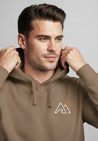 Young man adjusting hood of brown hoodie with white and orange mountain and sun logo on chest, light background.