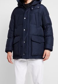 Giacca puffer blu navy con cappuccio, dotata di due tasche frontali e chiusure a scatto. Realizzata in tessuto strutturato e isolante per garantire calore.
