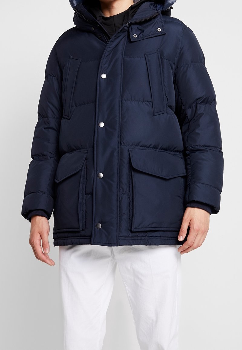 Giacca puffer blu navy con cappuccio, dotata di due tasche frontali e chiusure a scatto. Realizzata in tessuto strutturato e isolante per garantire calore.