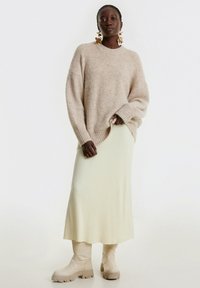 Beige gestrickter Pullover mit geripptem Muster, kombiniert mit einem blassgelben, gerippten Midirock und knielangen beigen Stiefeln. Goldene Blumenohrringe.