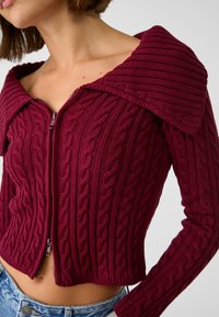Bordeauxkleurige cropped kabelgebreide trui met een opvallende geribbelde kraag en ritssluiting. Getextureerd patroon met aangesloten lange mouwen.