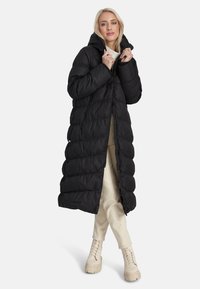 Naisterahvas, kes kannab pikka musta tikitud puffjope kapuutsiga, kreemivärvi pükse ja kreemivärvi nööpidega saapaid, seismas lihtsa tausta ees.