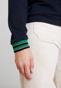 Námořnická mikina s proužkovanými manžetami v zelené, černé a zlaté barvě, doplněná světlými kalhotami. Pozornost věnována textuře látky a barvám.