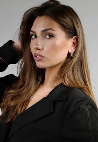 Pendientes de aro dorados, americana negra con solapas marcadas, textura suave, cabello largo castaño, maquillaje neutro sobre un fondo gris claro.