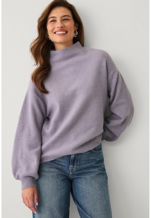 Smilende kvinde med bølget brunt hår iført lilla sweater med puffede ærmer og blå jeans, stående mod en hvid væg.