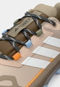 Κοντινό πλάνο σε μπεζ παπούτσι Adidas Terrex με μαύρα κορδόνια περασμένα μέσα από πολύχρωμες τρύπες και λευκές πλαϊνές ρίγες σε καφέ πλέγμα γλώσσας.