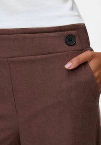 Bruine corduroy broek heeft een gladde textuur, een zijzak en een zwarte knopenaccent. De tailleband heeft een ribbelpatroon.