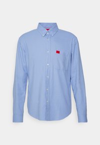 Camisa de manga larga azul claro hecha de algodón, con cuello abotonado, bolsillo frontal con una pequeña etiqueta roja y botones blancos.