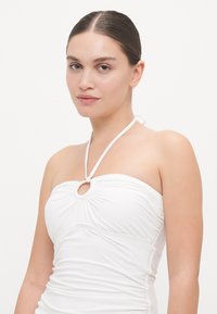 Witte halter-nekswimsuit met gathers, cirkelvormige uitsnede aan de voorkant en een gladde textuur. Slanke straps zijn als een strik achteraan.