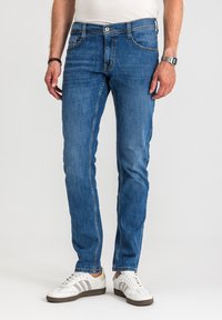 Mężczyzna w niebieskich dopasowanych dżinsach, białych trampkach z szarymi paskami i białej koszuli, stojący na jednolitym jasnym tle.