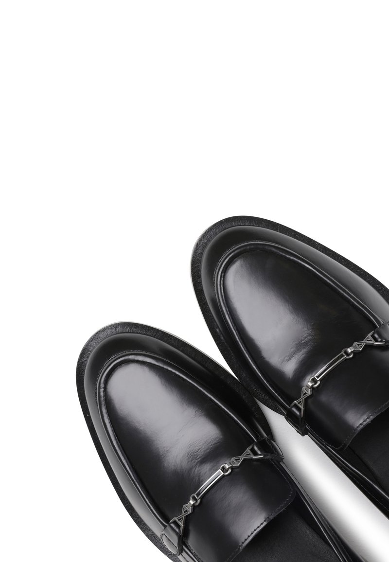 Des mocassins en cuir noir présentent une finition brillante, un bout arrondi et une chaîne décorative en métal argenté sur le cou-de-pied, reposant sur une semelle noire.