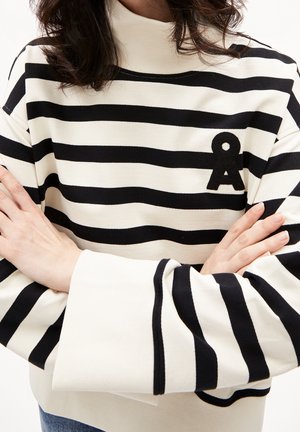 Henkilö pukeutuneena mustavalkoraidalliseen pooloneuleeseen, jossa rinnan kohdalla musta "Å"-logo, kädet ristissä.