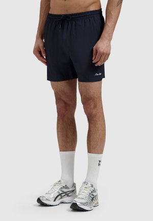 Mann trägt schwarze Shorts mit Kordelzug, weiße Kniesocken und silberne Laufschuhe und steht vor einem einfarbigen Hintergrund.