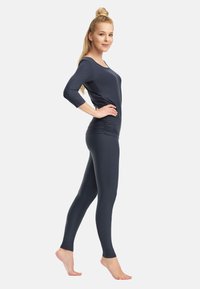 Pikendatud blondidega naine, kes kannab tihedat tumehallist pika varrukaga toppi ja retuuse, seisab paljajalu külgvaates.