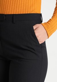 Mustat housut, joissa on mukautettu istuvuus, litteä etuosa, sivutaskut ja sileä kangas, yhdistettynä ribbikuvioiseen oranssiin yläosaan.