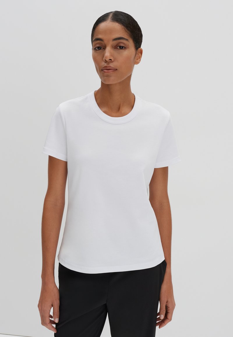 Weißes Baumwoll-T-Shirt mit rundem Halsausschnitt, kurzen Ärmeln und lockerer Passform, kombiniert mit schwarzen Hosen. Glatte Textur, keine sichtbaren Muster.