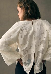 Blouse blanche ample en tissu transparent avec un motif floral, présentant des manches larges et un détail de fente au dos.