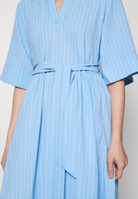 Ljusblå klänning av mjukt tyg med vita vertikala ränder. Har en V-ringning, vida ärmar och ett tygbälte för att cincha midjan.